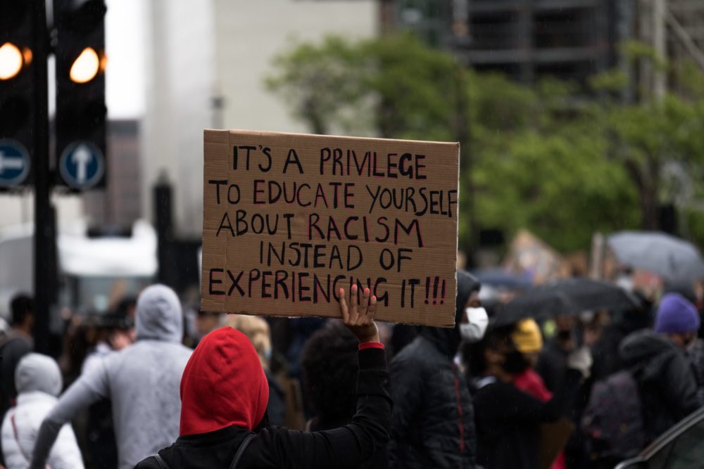 protester holds cardboard handmade sign that reads, "It's a privilege to educate yourself about racism instead of experiencing it."