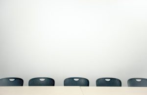 alt text: image is a color photograph of classroom chairs pushed up against a desk; title card for Amy Evan's creative nonfiction piece "The Stoics"
