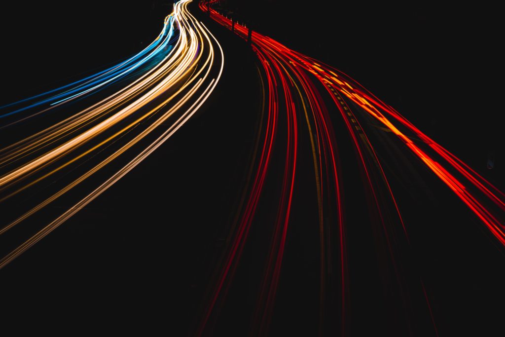alt text: image is a color photograph of a streak of car lights at night; title card for Myna Chang's creative nonfiction piece "We Were the Wild Hunt"