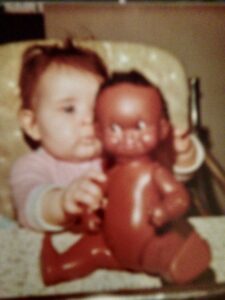 Image is a photograph of a young baby in a high chair with a doll.