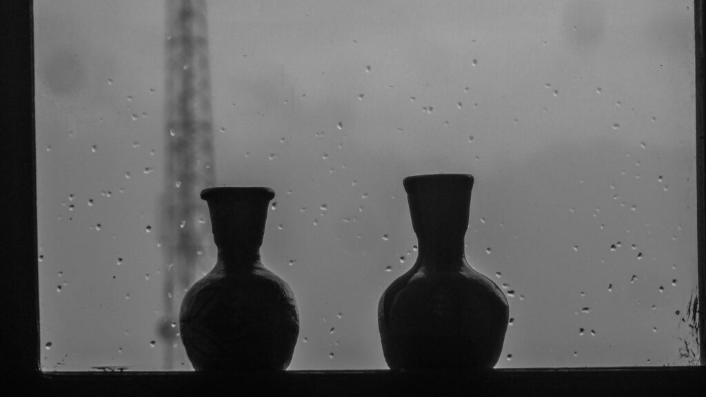 Image is a black-and-white photograph of two urns in front of a rain-spattered window; title card for the new flash fiction, “Particulate Matter,” by Rigel Olivera.